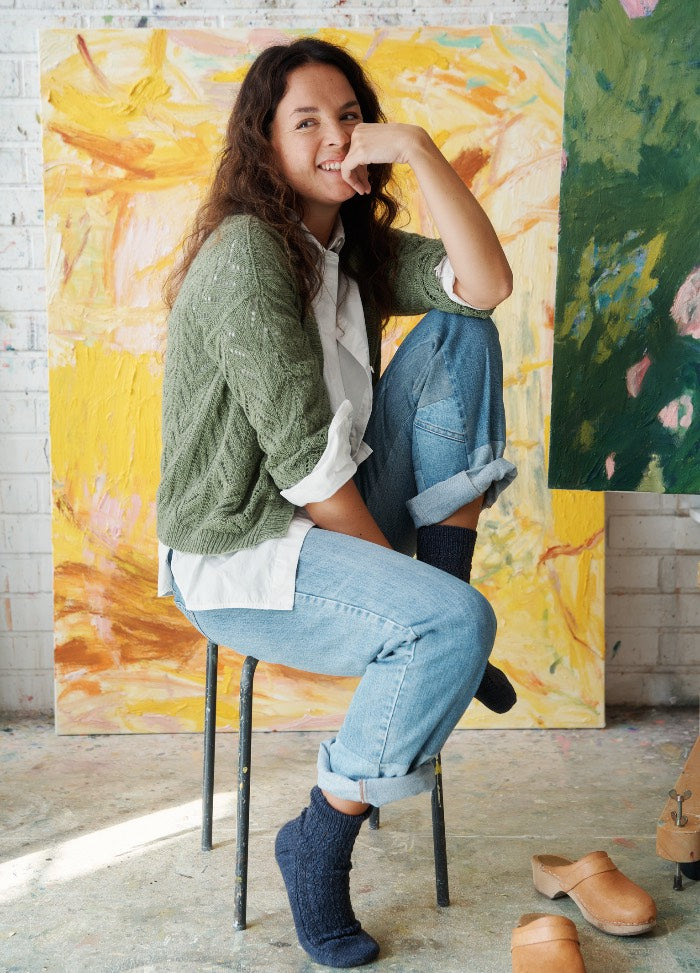 Thuja Cardigan: model seated in green knit cardigan, white shirt, and jeans, with abstract art in the background.