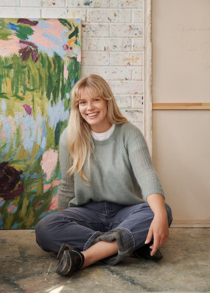 Selsey Sweater: light green knit sweater worn with dark jeans and black shoes, seated in front of abstract art.