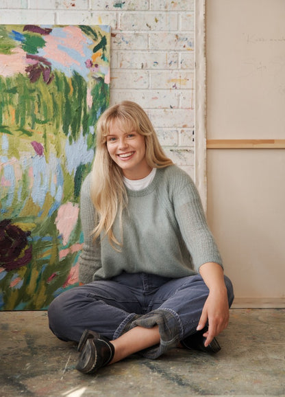 Selsey Sweater: light green knit sweater worn with dark jeans and black shoes, seated in front of abstract art.