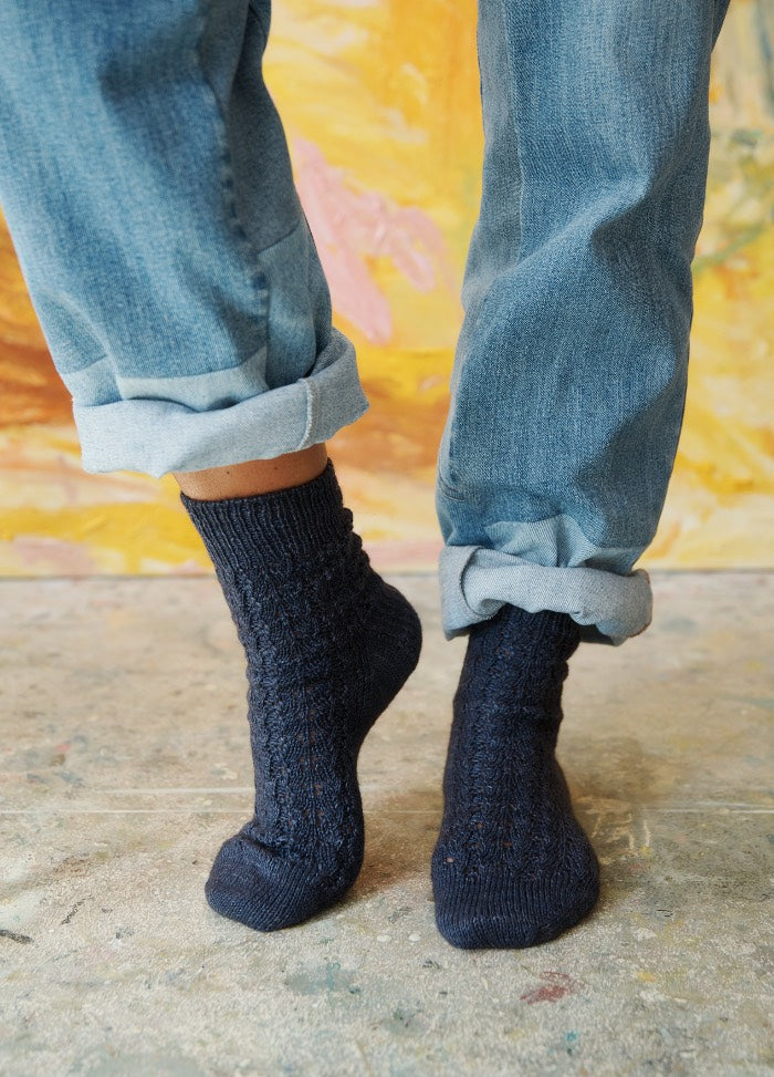 Blue Hour Socks: model in blue knit socks and rolled-up jeans, standing on a paint-splattered concrete floor.