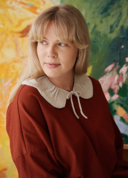 Flores Collar: white knitted bow-tied collar worn over a rust sweater, with abstract art in the background.