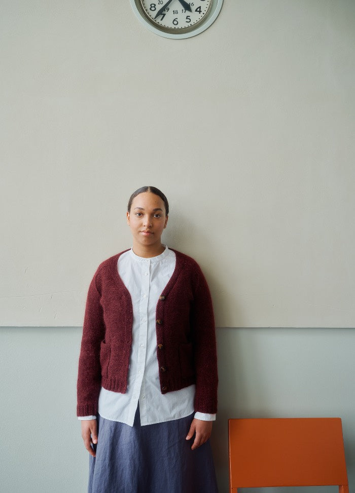 Maroon cardigan worn over a white shirt and blue skirt, with an orange chair and wall clock in the background.