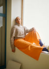 Oversized light-colored sweater paired with bright orange wide-leg pants, worn while sitting on a windowsill.