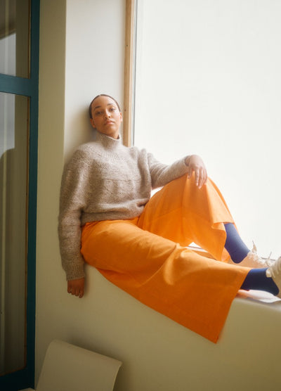 Oversized light-colored sweater paired with bright orange wide-leg pants, worn while sitting on a windowsill.