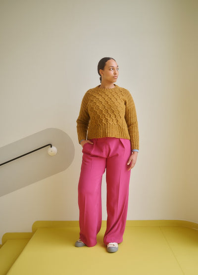 Mustard yellow cable-knit sweater paired with bright pink wide-leg pants, set against a minimalist indoor background.