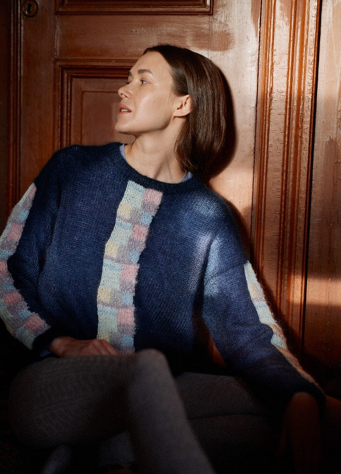 Dark blue sweater with pastel checkered patterns on the front and sleeves, shown seated against a wooden door.