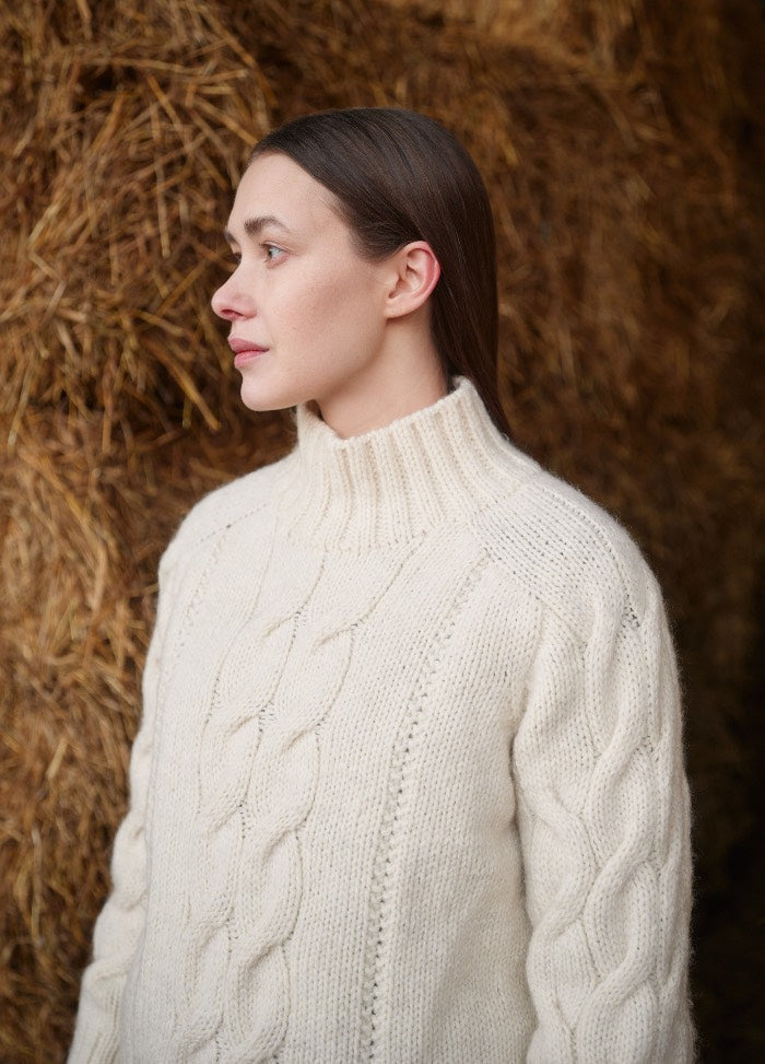 White cable-knit sweater with a high collar, shown against a rustic hay background.