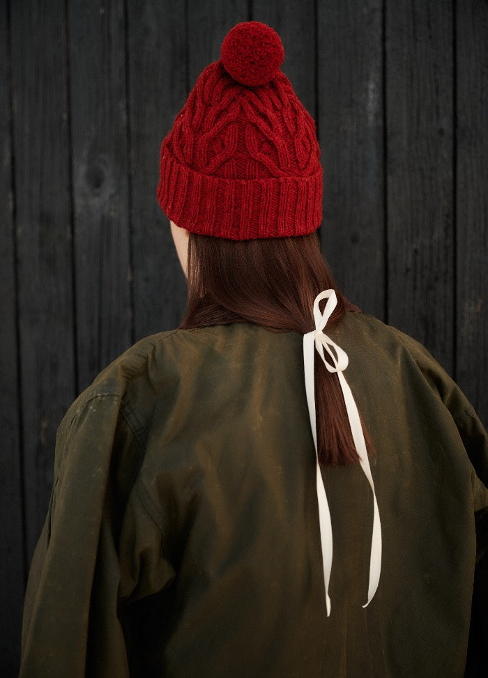 Red knitted hat with pom-pom, shown from the back with long hair and an olive jacket against black wooden planks.