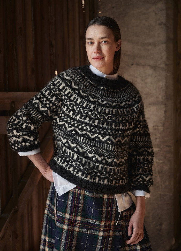 Black and white patterned sweater layered over a white shirt, paired with a colorful plaid skirt in an indoor setting.