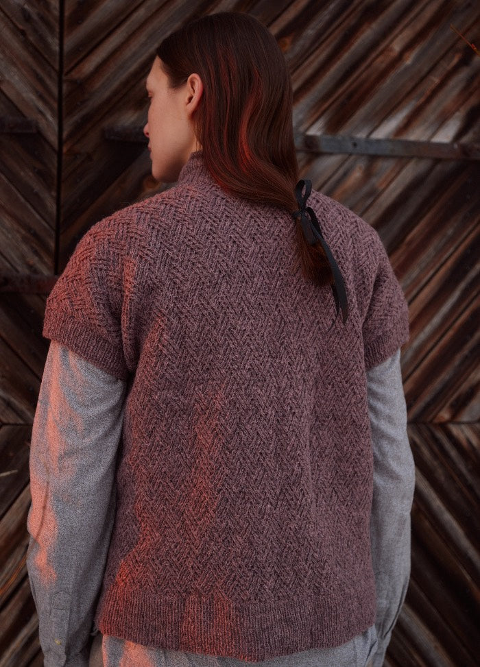 Earth-toned textured cardigan shown from the back, with a ribbon-tied hairstyle against a wooden plank background.