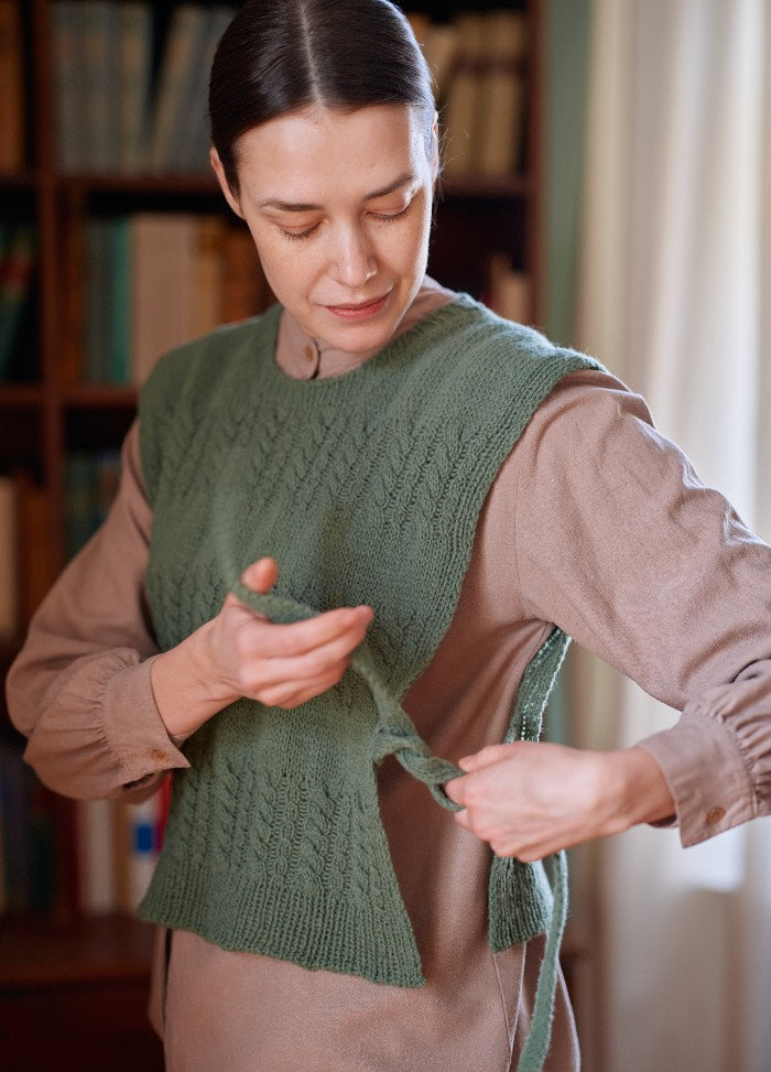 Green knitted vest styled over a beige shirt, shown being adjusted at the side in a cozy room with bookshelves.