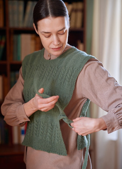 Green knitted vest styled over a beige shirt, shown being adjusted at the side in a cozy room with bookshelves.