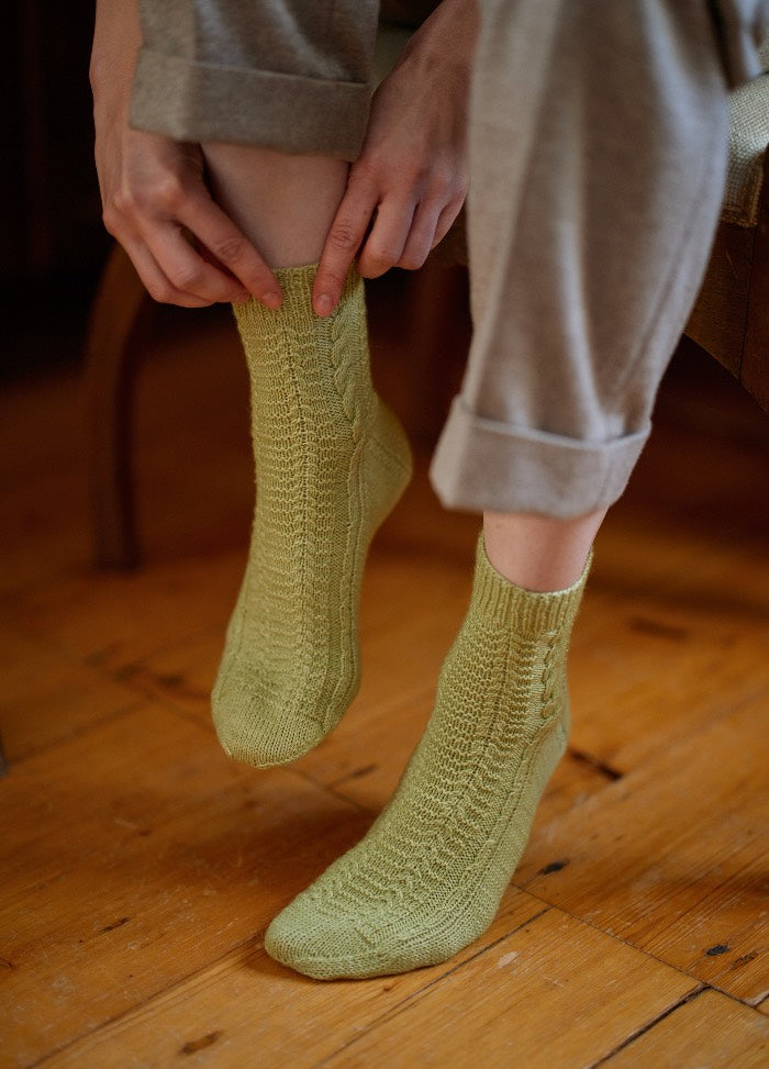 Light green knitted socks being adjusted while seated on a wooden floor, paired with rolled-up light pants.