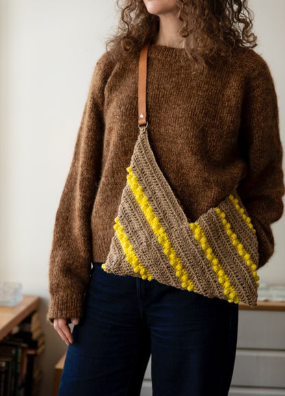 Mimosa Bag: knitted shoulder bag with diagonal yellow stripes and a leather strap, worn with a brown sweater and dark jeans.