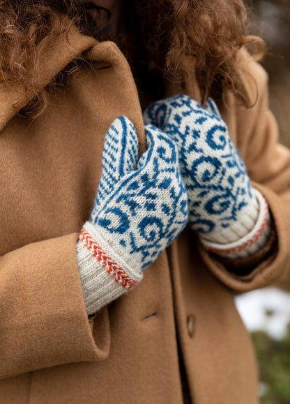 Akantus Mittens: blue and white patterned knitted mittens with red decorative stitching near the wrist, worn with a brown coat outdoors.