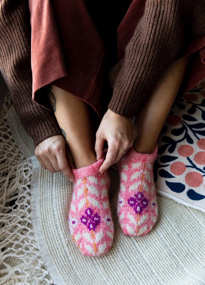 Juni Slippers: pink knitted slippers with purple and white patterns, worn on a woven rug with decorative mat accents.