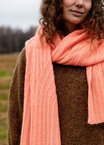 Plot Twist Scarf: pink knitted scarf wrapped around the neck, worn with a brown sweater in an outdoor setting.