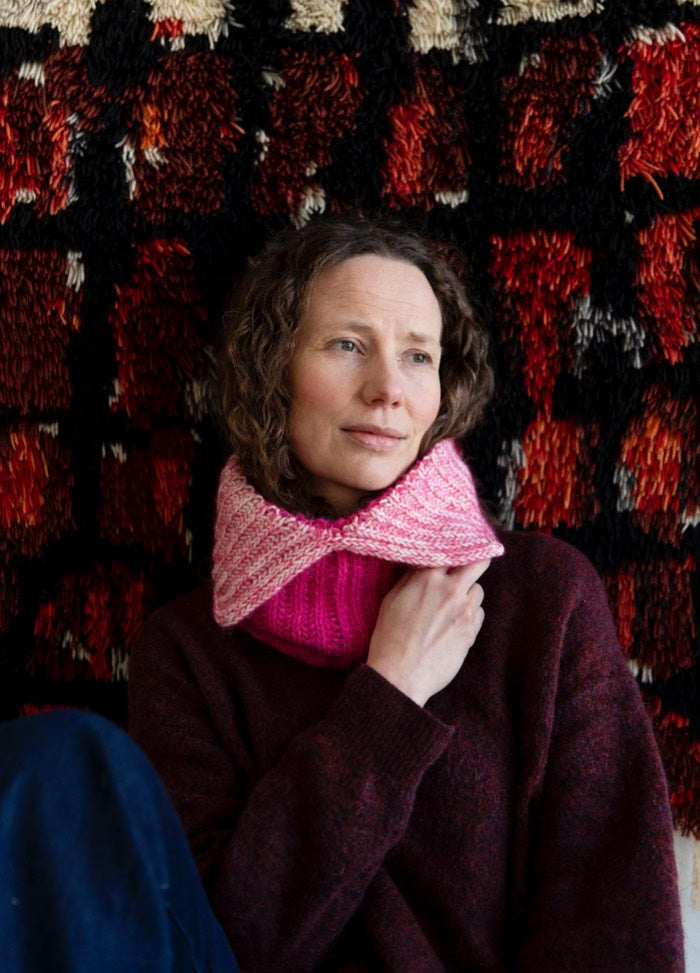 Kitty Cowl: pink knitted cowl with a textured pattern, worn with a dark sweater against a red and black textile background.