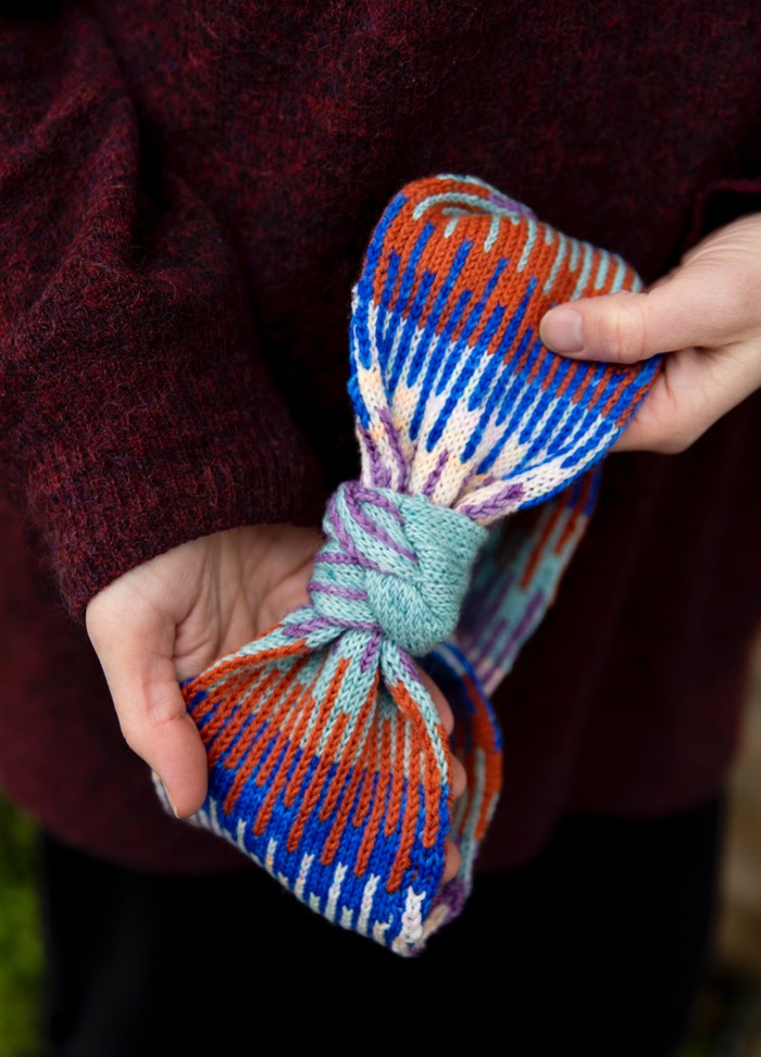 Between the Lines Headband: multicolored knitted headband with a bow-like tie in light green, held by a person in a dark red sweater.