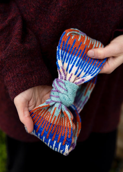 Between the Lines Headband: multicolored knitted headband with a bow-like tie in light green, held by a person in a dark red sweater.