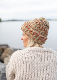 Halla Beanie: knitted beanie worn with a cream sweater, photographed from behind near water and rocks.