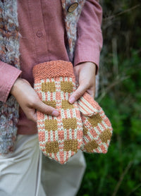 Outside the Box Mittens: checkered knit mittens in orange and beige, held outdoors with pink shirt and textured vest.