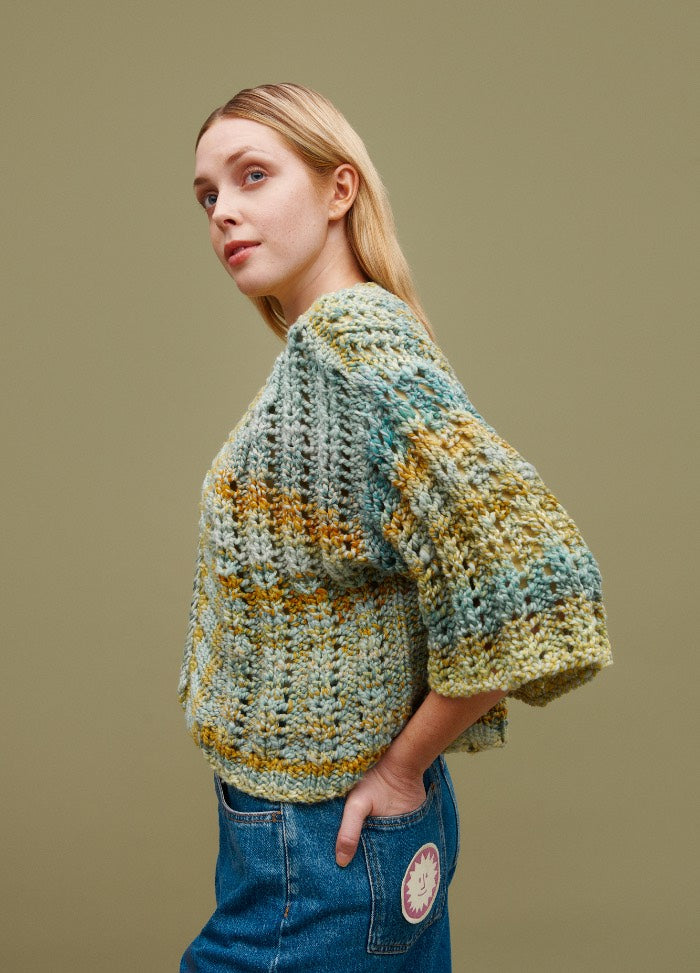 Petit Palais Cardigan: multicolored textured knit in green, blue, and yellow, styled with blue jeans against an olive background.