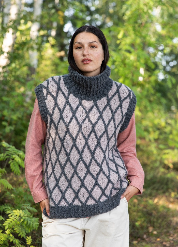 Jorvin Vest: light knit vest with dark diamond pattern and ribbed collar, layered over a pink shirt in a natural outdoor setting.