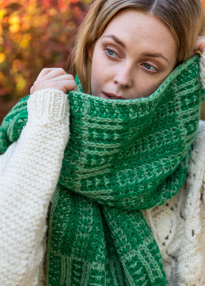 Vice Versa Scarf: green knit scarf with lighter green stripes, styled with a cream sweater in an autumn outdoor setting.
