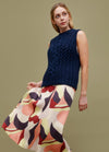 Cable Blur Vest: dark blue cable-knit sleeveless vest styled with a colorful abstract skirt against a muted green background.