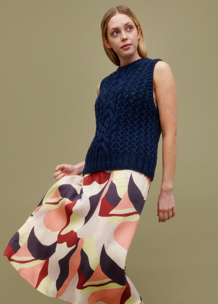 Cable Blur Vest: dark blue cable-knit sleeveless vest styled with a colorful abstract skirt against a muted green background.