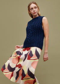 Cable Blur Vest: dark blue cable-knit sleeveless vest styled with a colorful abstract skirt against a muted green background.
