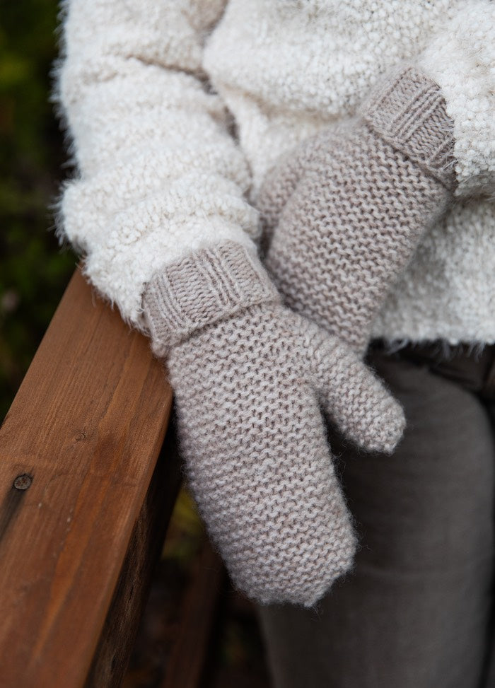 Inkeri Mittens: light gray textured knit mittens worn with a white fluffy sweater, hands resting on a wooden surface.