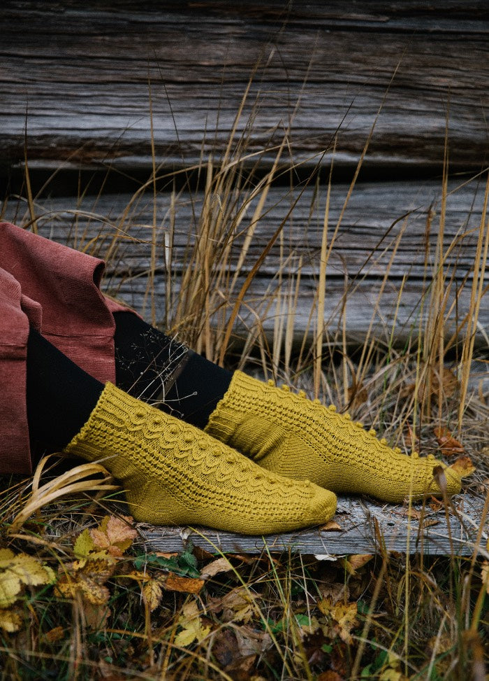 Craspedia Socks: mustard yellow textured socks worn with black leggings and pink pants, seated on dry grass and wooden planks