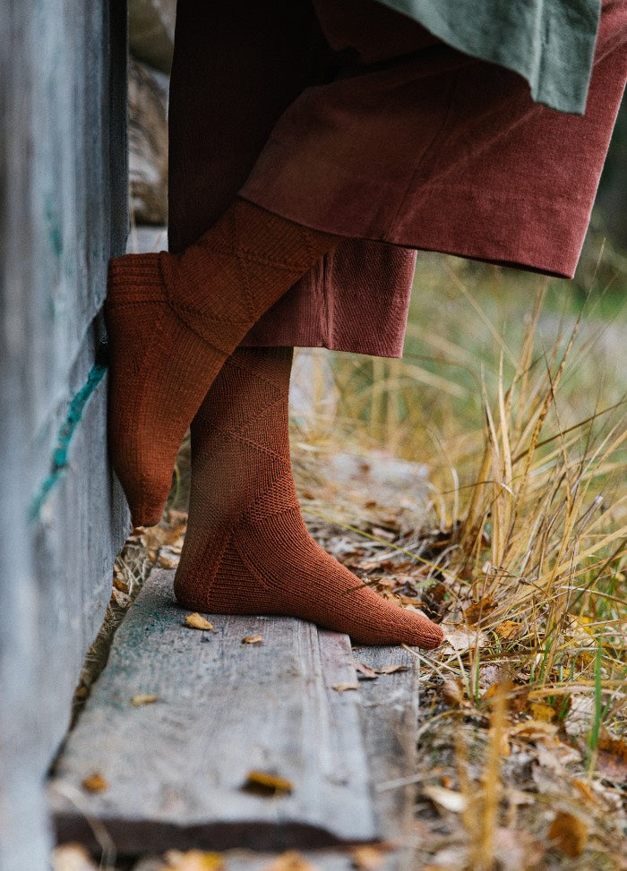 Bembe Socks: rust-colored socks with subtle pattern, worn with rolled-up pants, standing on wooden surface near dry grass and leaves