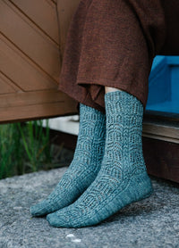 Branches Socks: teal-colored knitted socks with intricate pattern, worn by seated person near stone surface and wooden door
