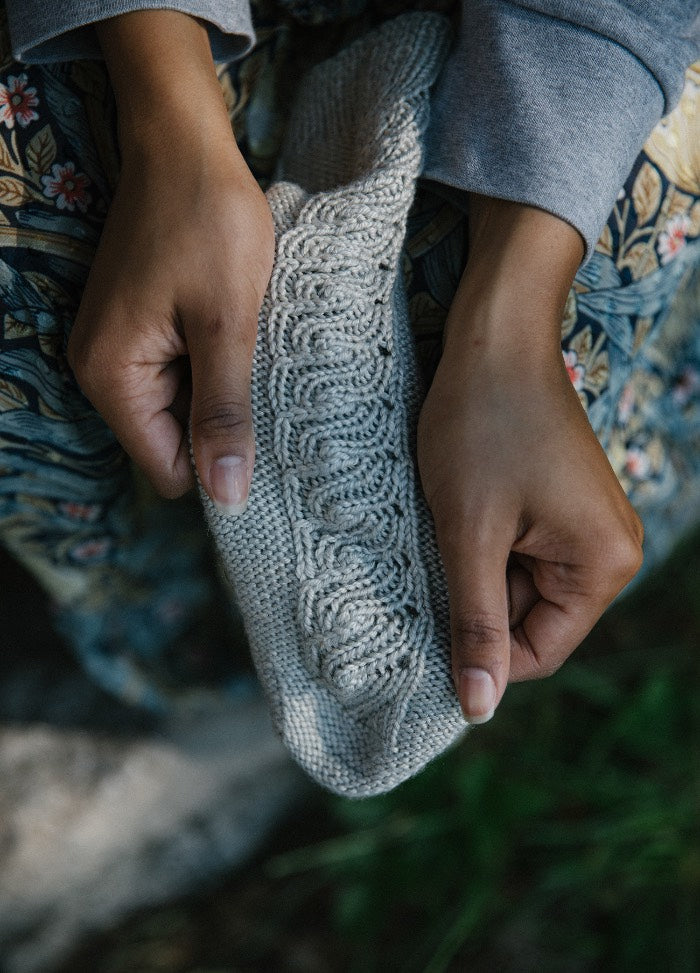 Riverbed Socks: light gray sock with intricate cable knit pattern, held in hands over floral-patterned fabric