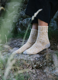 Lazy Daisy Socks: light-colored speckled socks with yellow and gray flecks, worn with rolled-up black pants on rocky ground outdoors