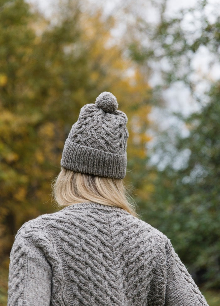 Lempi Hat: cable knit hat with pom-pom, shown from back with matching sweater and autumn foliage.
