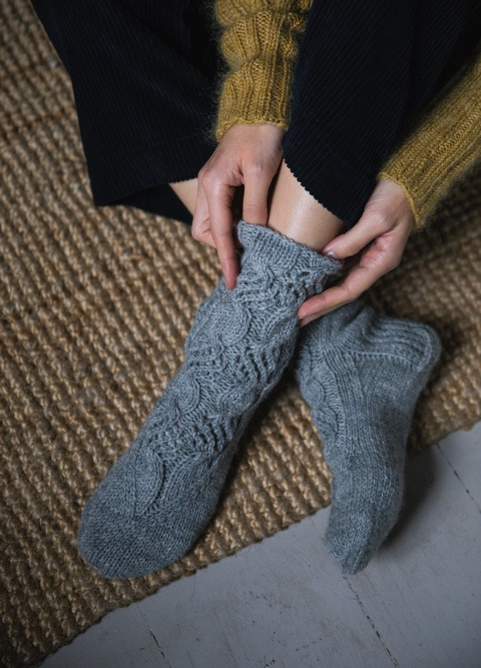 Mielenrauha Socks: grey knit with intricate pattern, worn seated on woven rug with yellow sweater.