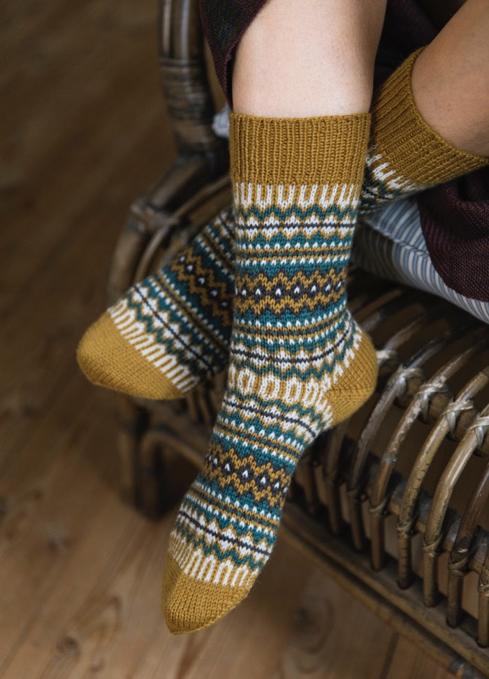 Taipale Socks: mustard yellow knit with multicolor geometric patterns, worn seated on wicker chair.
