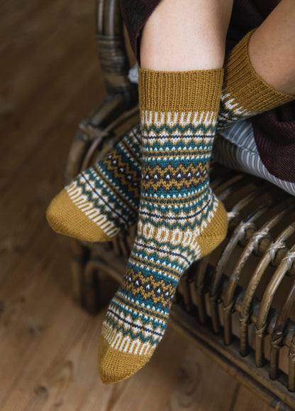 Taipale Socks: mustard yellow knit with multicolor geometric patterns, worn seated on wicker chair.