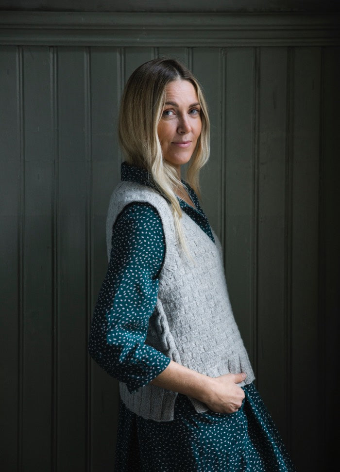 Vakka Vest: grey knit worn over dark green polka dot dress, shown against wooden panel backdrop.