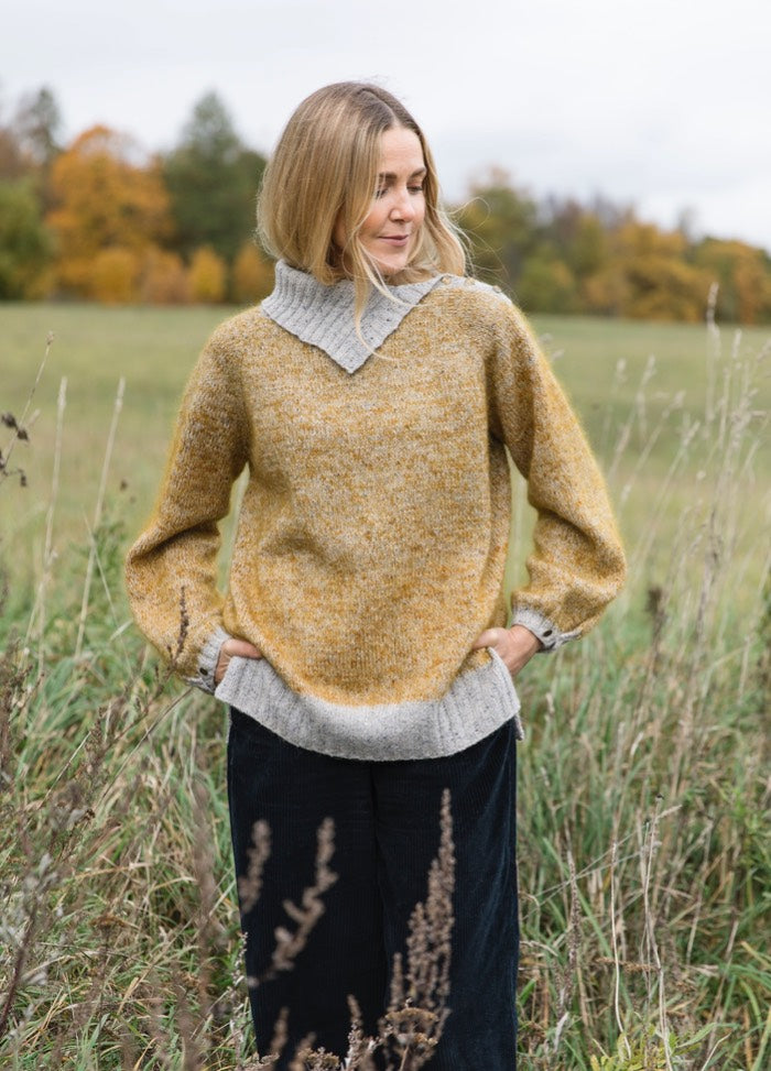 Malva Sweater: yellow knit with grey trim, worn outdoors among tall grass and autumn trees.