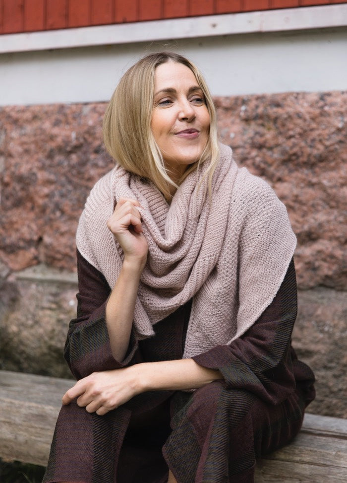 Usva Shawl: knitted wrap draped over shoulders, shown seated by stone wall and red building.