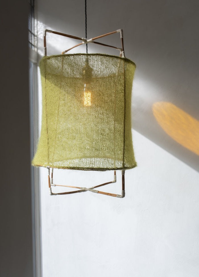 A hanging lamp with a soft, green fabric shade and thin metal frame casts a warm glow on a white wall, creating a calm and minimalist ambiance.