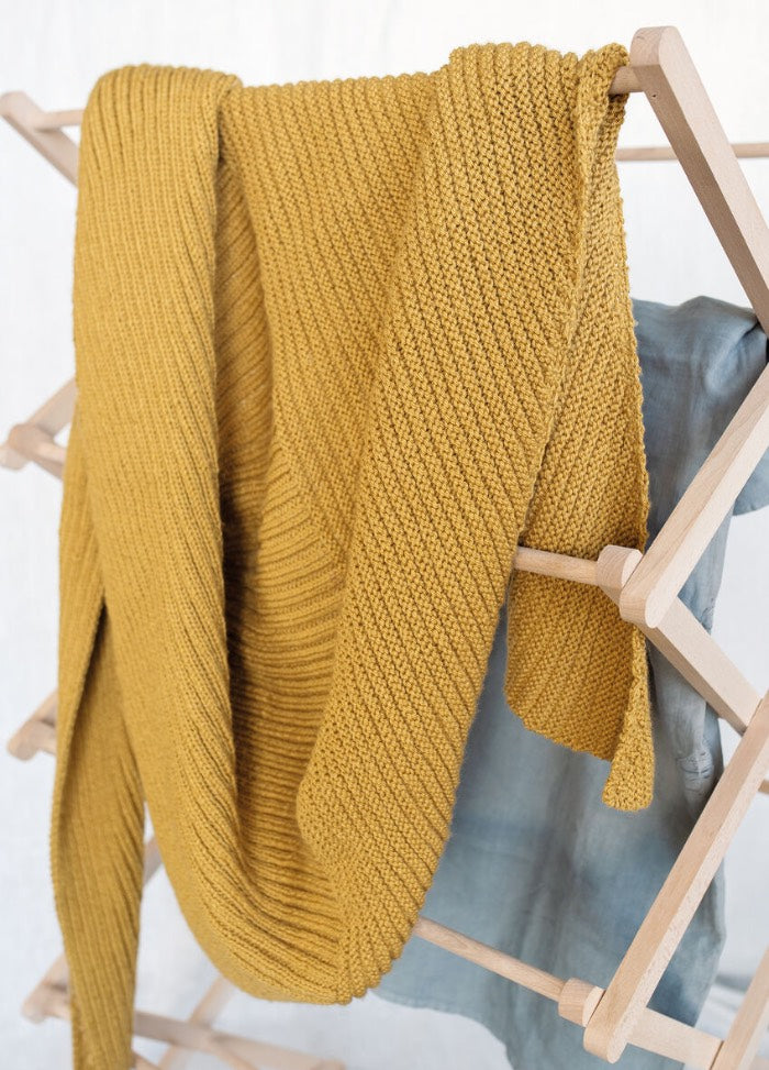 Mustard yellow knit blanket draped over a wooden drying rack, with a soft blue-gray fabric beneath it. The scene feels cozy and inviting.