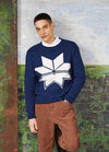 A man stands against a textured wall, wearing a blue sweater with a large white geometric star and brown corduroy pants, conveying a casual tone.