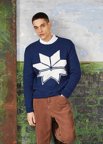 A man stands against a textured wall, wearing a blue sweater with a large white geometric star and brown corduroy pants, conveying a casual tone.