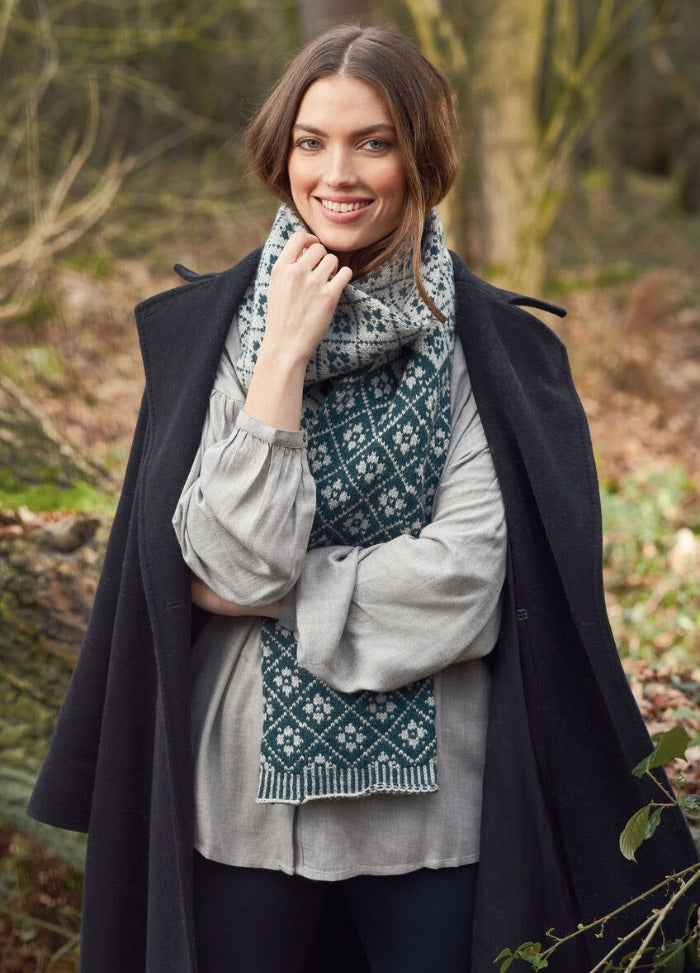 Woman smiles in a forest wearing a patterned scarf, light gray blouse, and dark coat. The scene feels cozy and serene, with soft autumn colors.